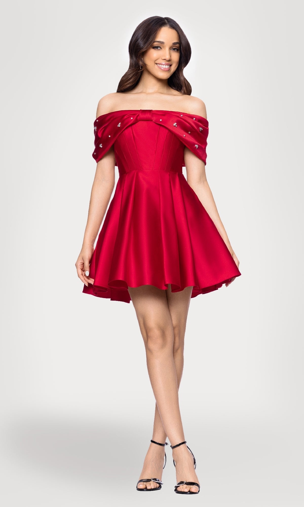 A woman smiles in the Off-Shoulder Red Hoco Dress: Blondie Nites 5695BN, featuring a corset-style bodice and flared skirt, paired with black strappy heels against a plain light background.